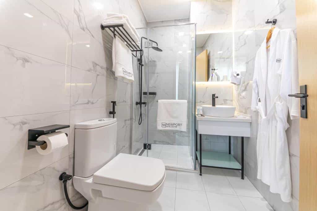 Modern hotel bathroom with marble-style tiles, glass shower, white vanity, and hanging bathrobes.
