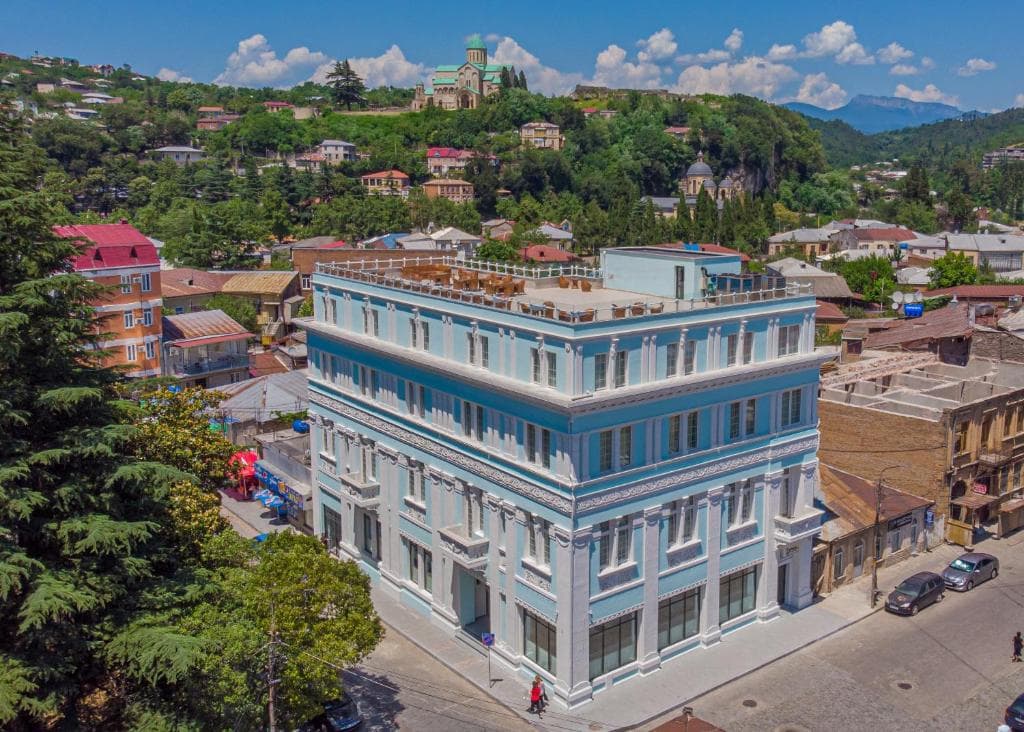 Light blue hotel building with a rooftop terrace in a hillside city with a prominent cathedral in the background.