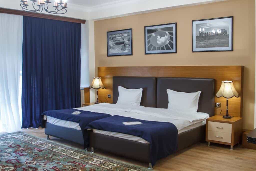 Hotel room with two twin beds, dark blue runners, bedside lamps, and framed wall art.