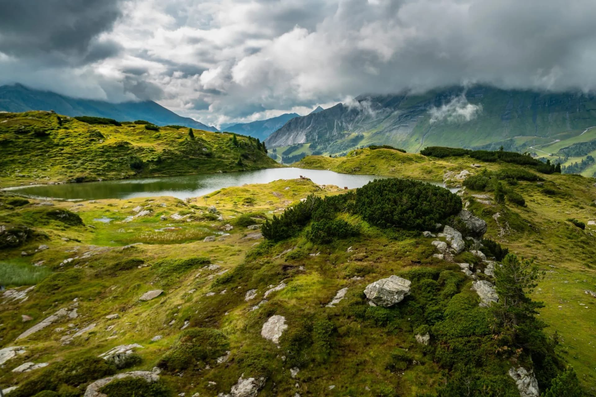 Alpine tarn surrounded by green hills and dramatic mountains under cloudy skies in Obertauern.