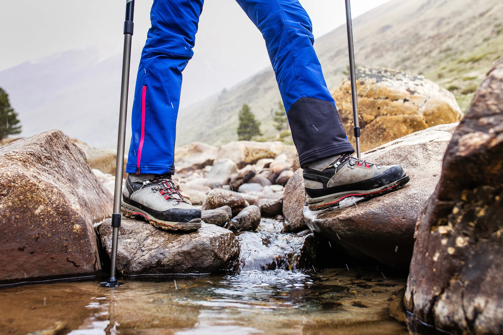 Hiking boots, waterproof overtrousers and poles crossing a rocky mountain stream