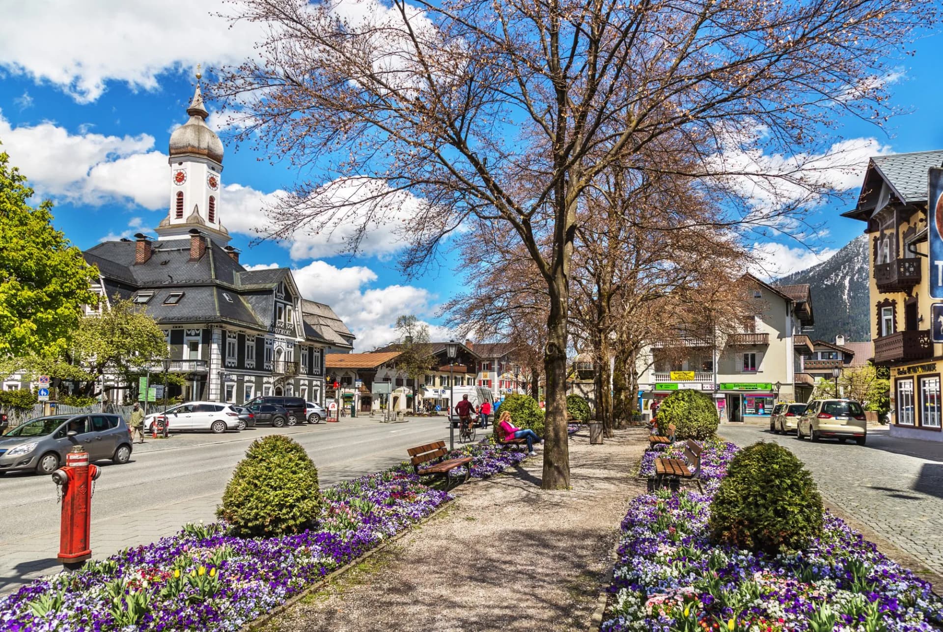 Street in Garmisch-Partenkirchen with church tower, flower beds, and snow-capped mountain backdrop.
