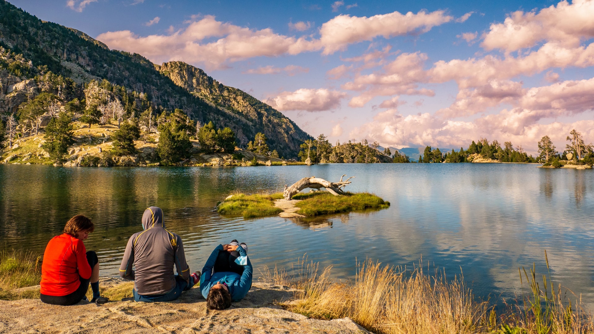 Three people sitting and relaxing by an alpine lake surrounded by mountains and pine trees.