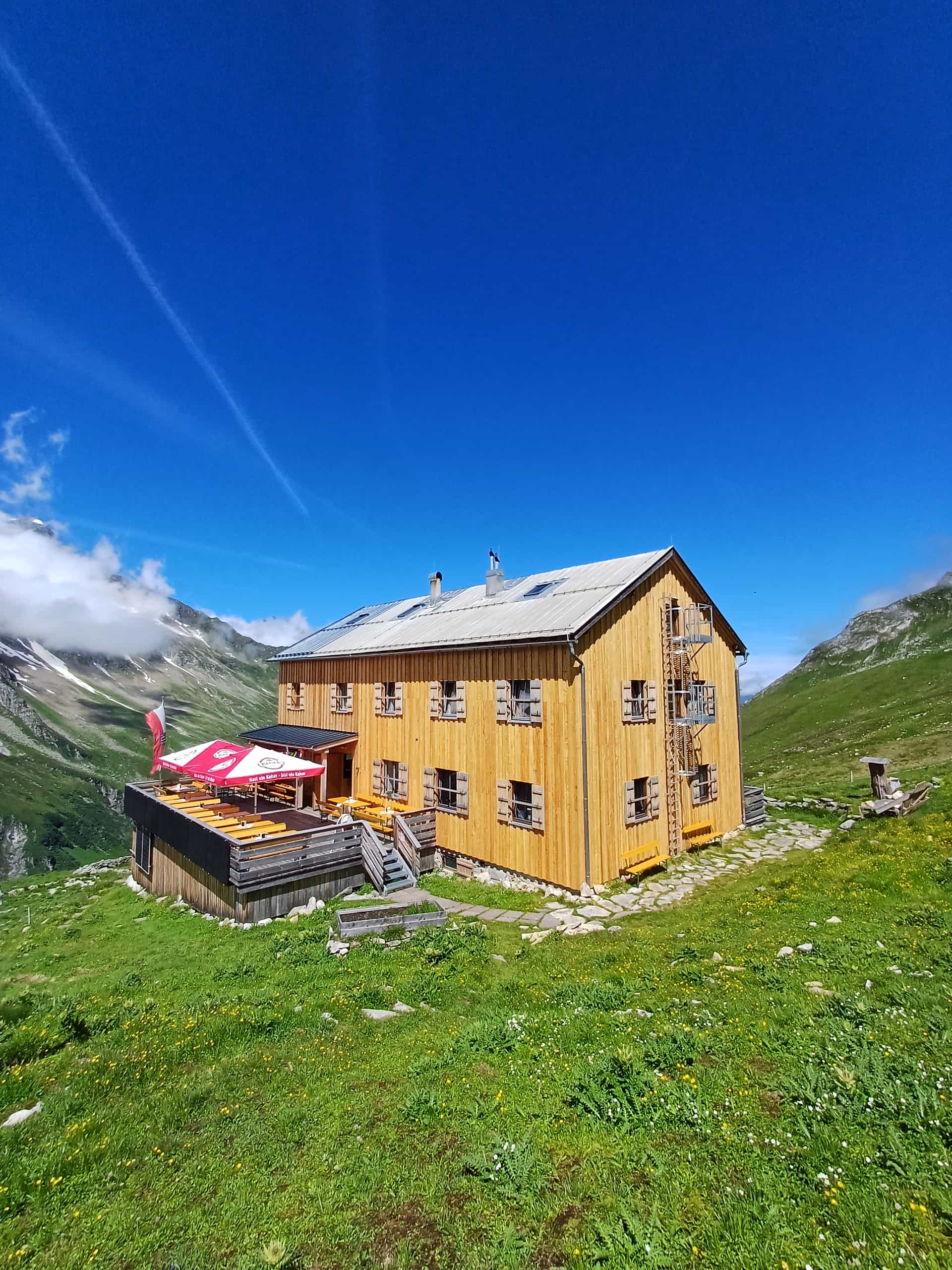 Wooden mountain hut with outdoor seating terrace in the Alps under a clear blue sky.
