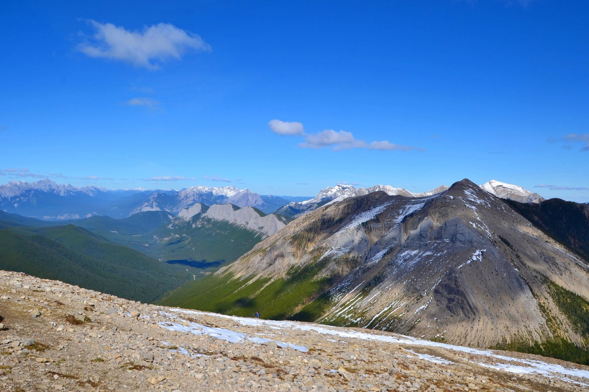 A bare stony ridge in the foreground looking across to a snow-streaked peak, with a green valley below and ranges beyond.