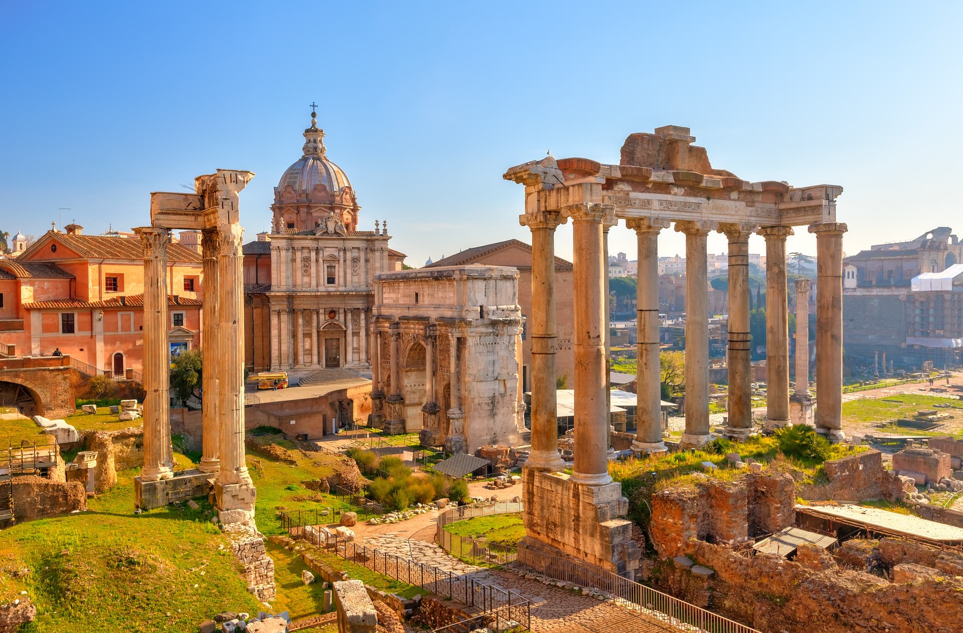 Ancient Roman Forum ruins with Temple of Saturn columns and church dome under clear blue sky in Rome.
