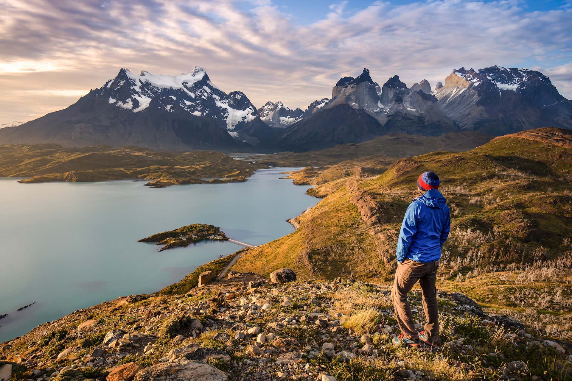 Hiker overlooking glacial lake and jagged, snow-capped mountains at sunset in Patagonia.