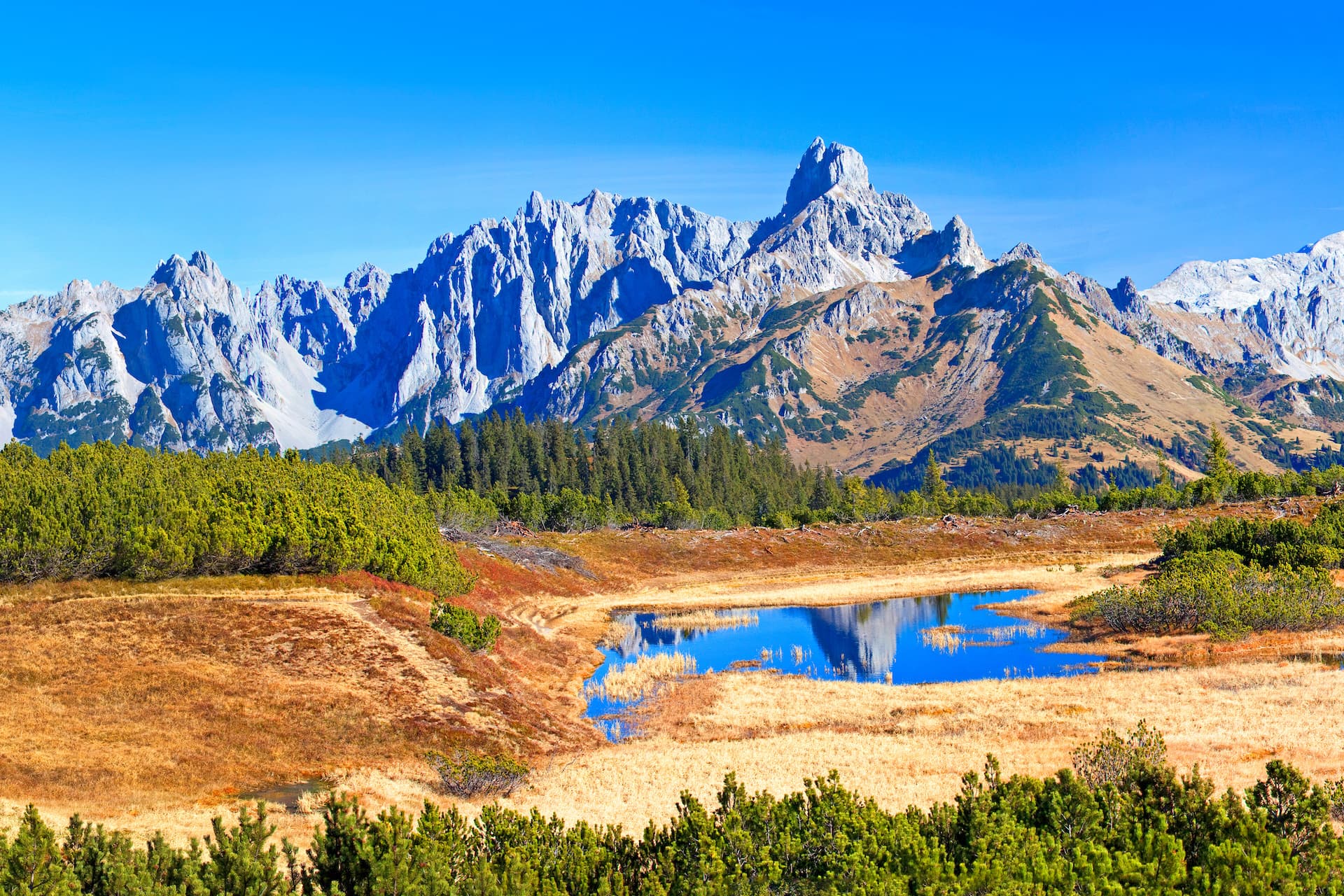Alpine tarn reflecting jagged, rocky mountains under a clear blue sky in autumn.