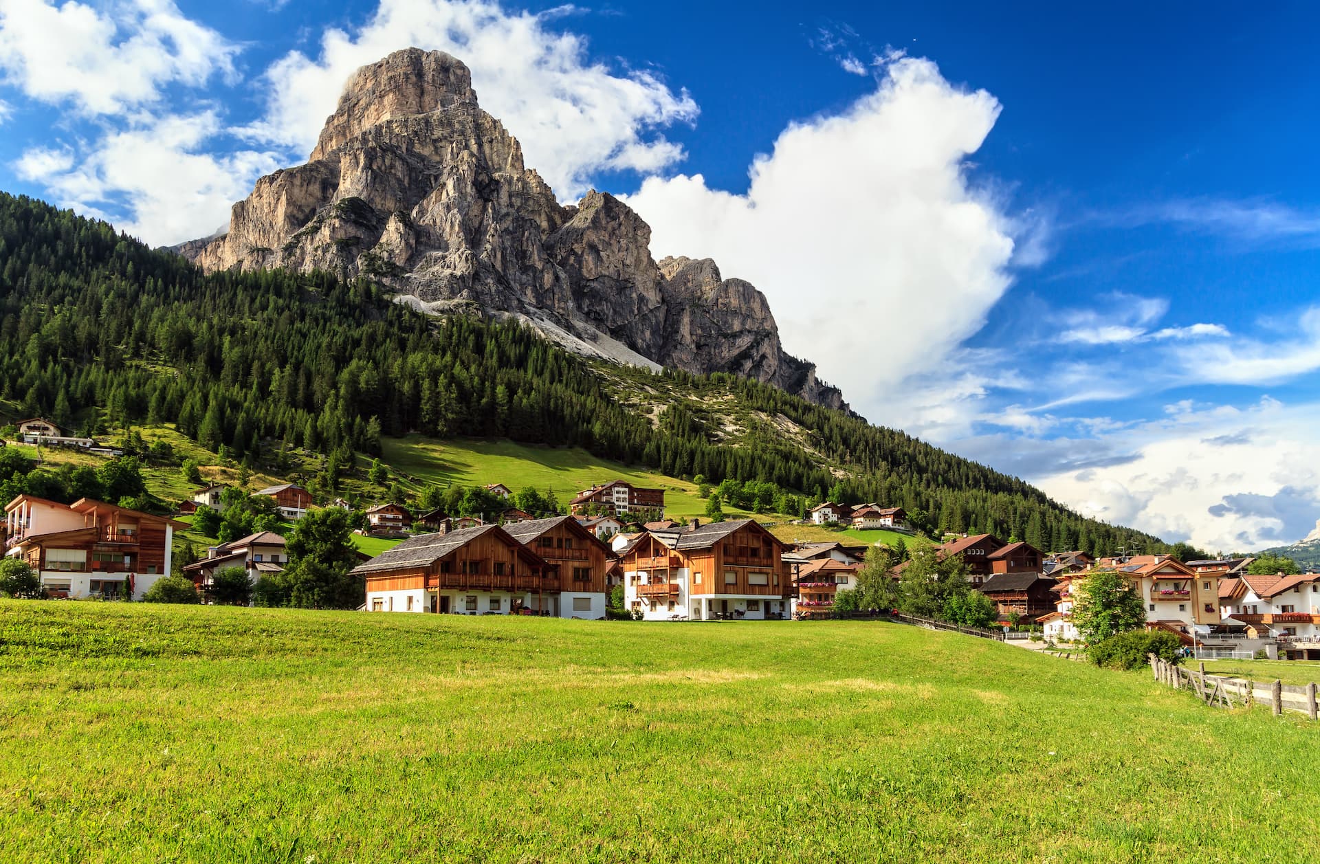 Alpine village with wooden chalets nestled below a massive rocky mountain under a blue sky.