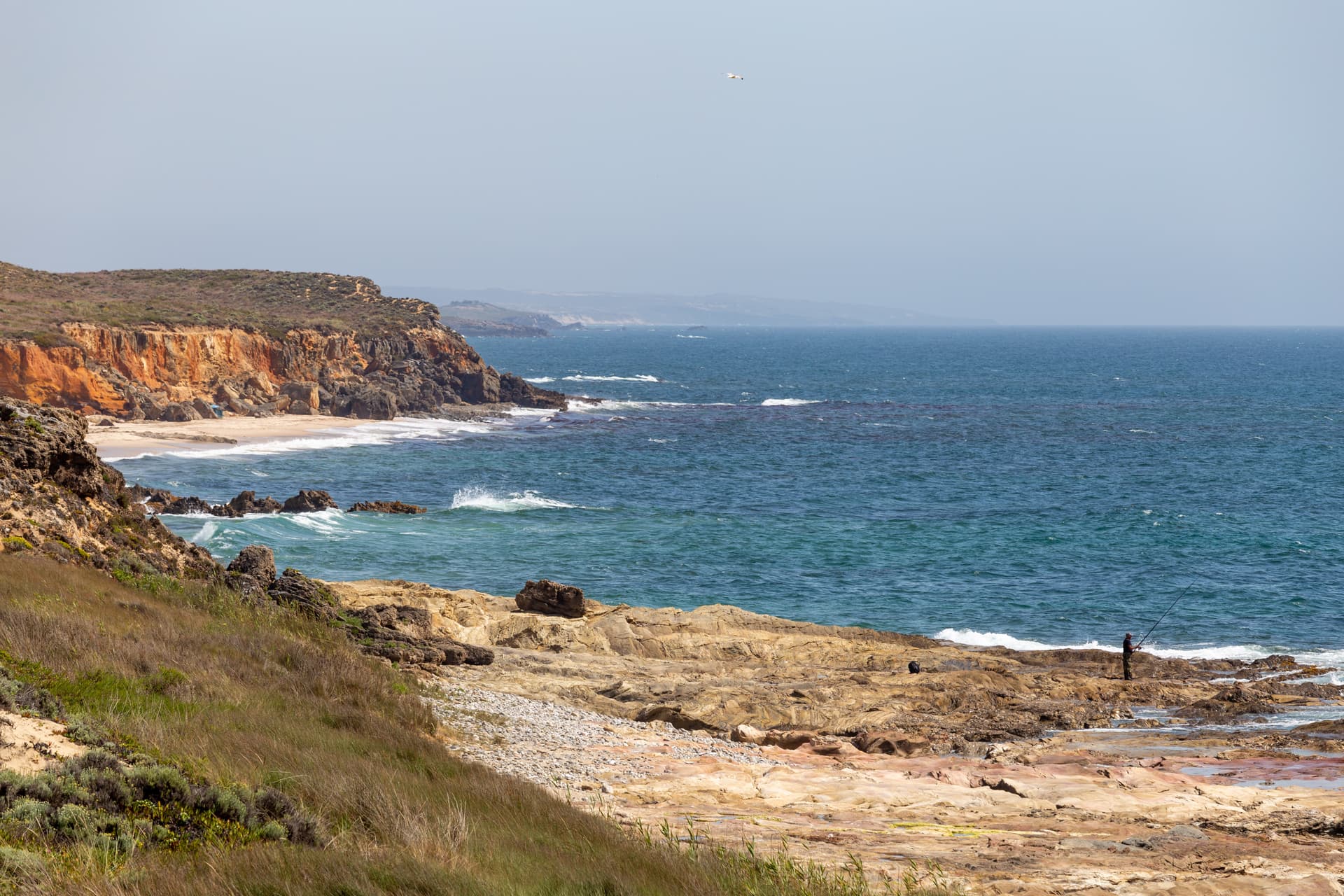 Fisherman on rocky shore next to blue ocean waves beneath orange cliffs at São Torpes beach.