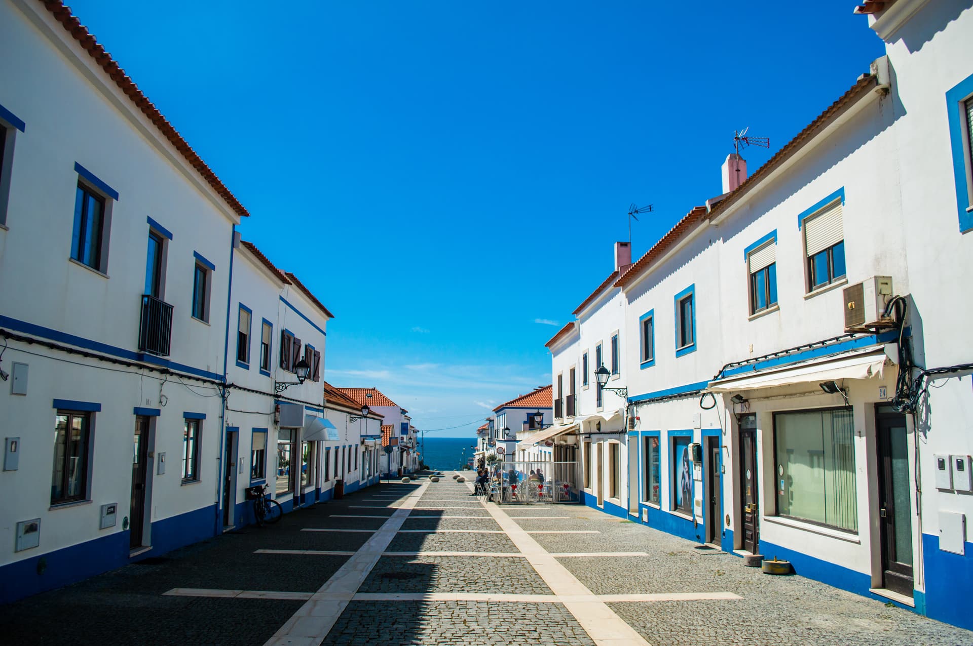 Cobblestone street leading to the sea between white and blue buildings in Porto Covo.