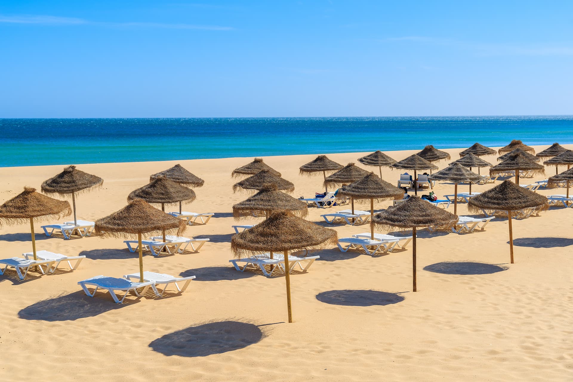 Straw umbrellas and white sunbeds on golden sand beach facing turquoise sea in Salema, Portugal.
