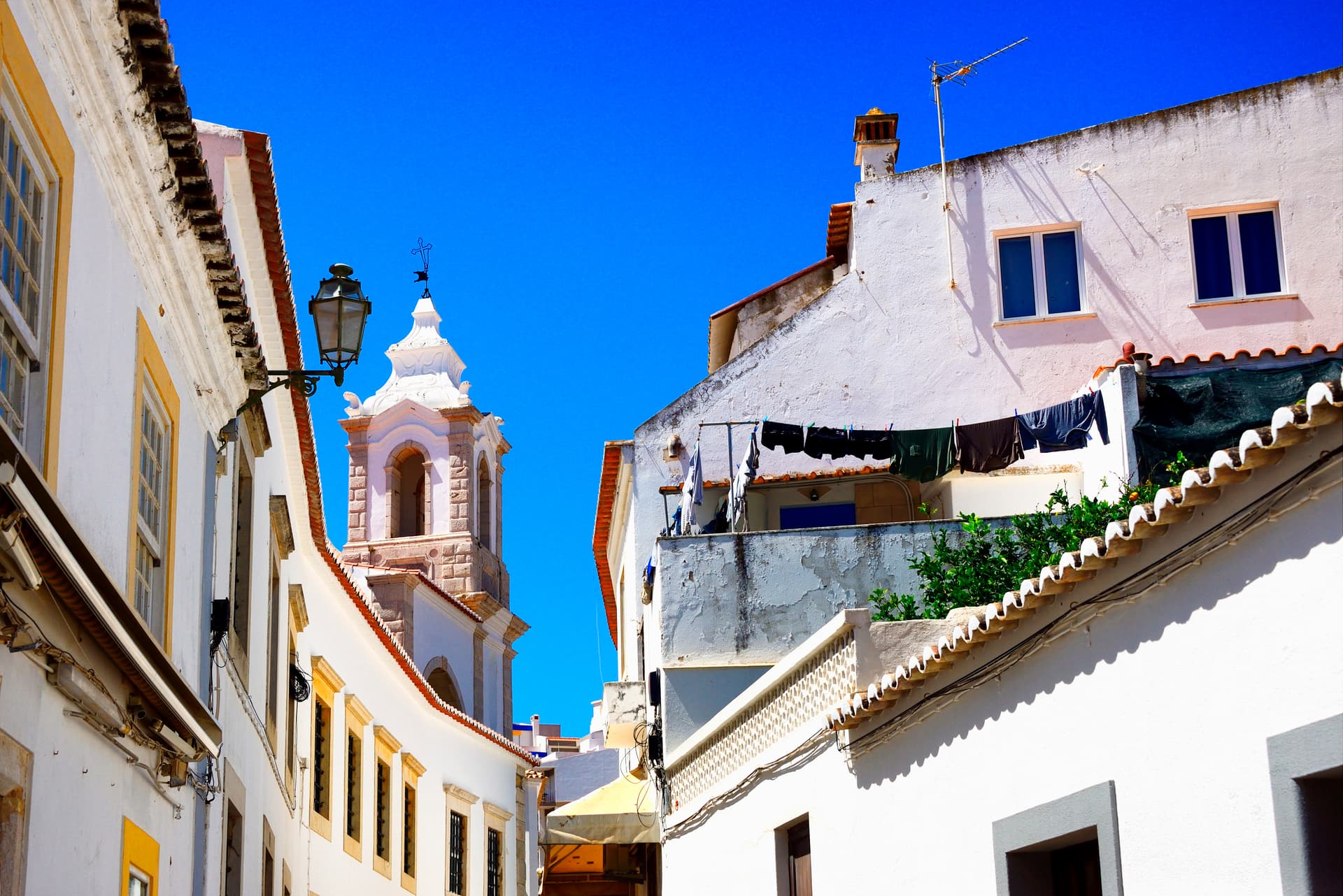 Narrow street in historic Lagos, Algarve, Portugal, with white buildings and a church tower under a bright blue sky.