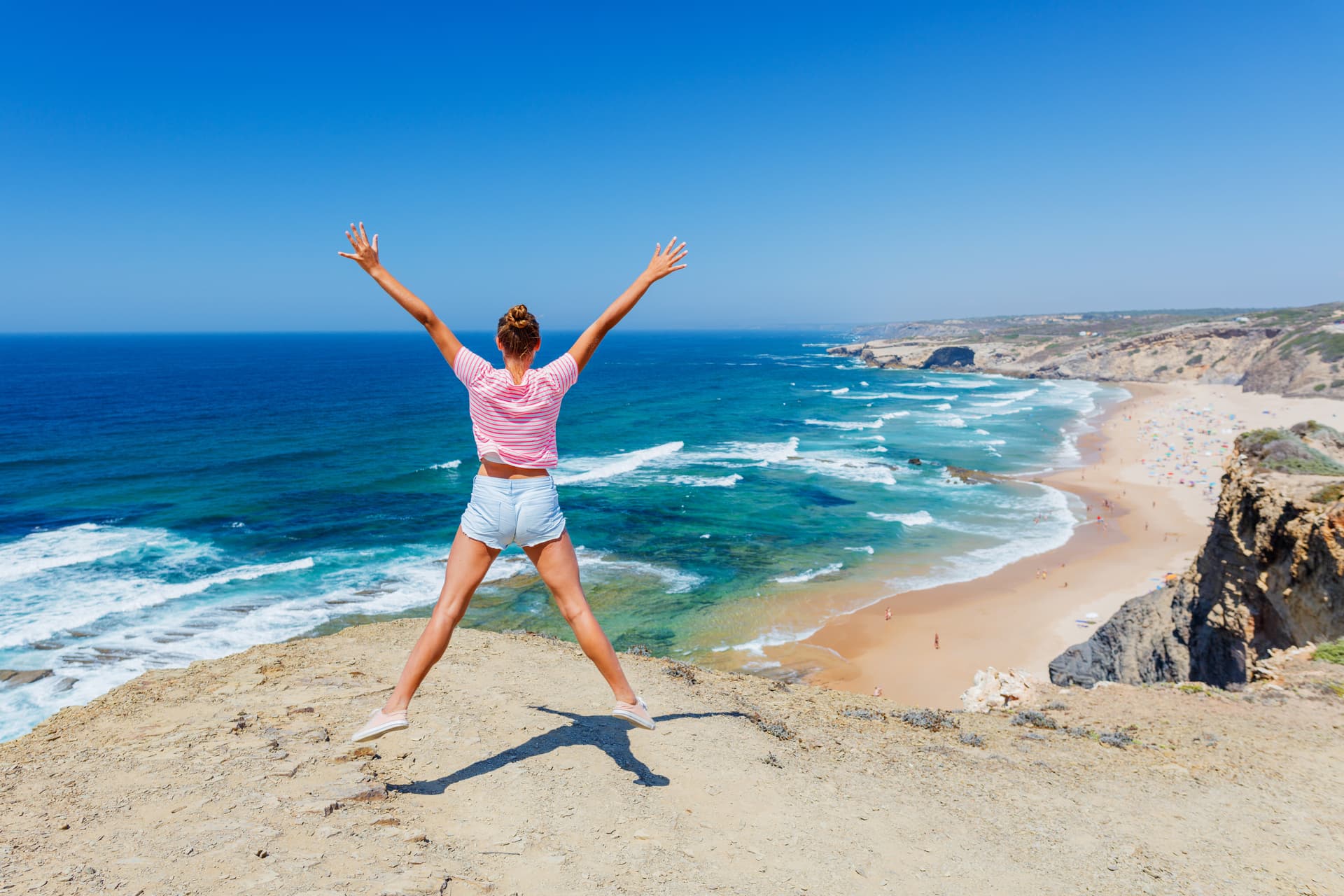 Woman jumping on cliff overlooking Lagos Algarve Coast beach and turquoise ocean waves.