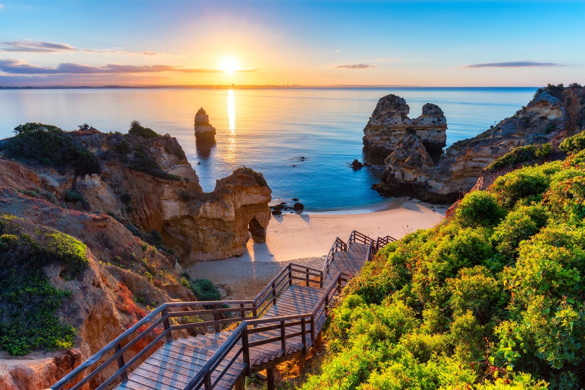 Wooden stairs lead to Camilo Beach with sea stacks at sunrise in Lagos, Algarve, Portugal.