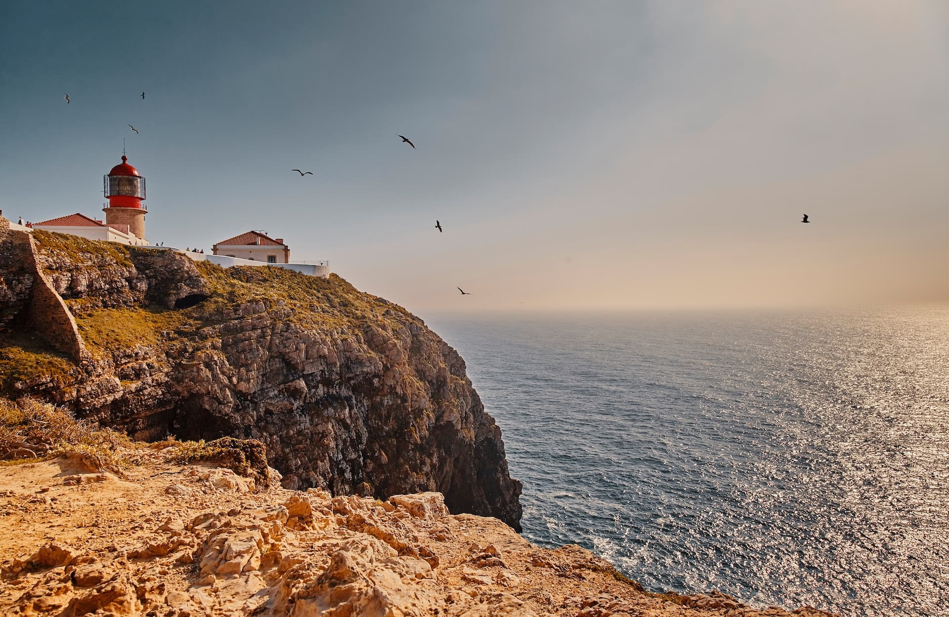 Lighthouse on cliff edge overlooking sparkling ocean with birds flying, Cape Saint Vincent, Portugal.