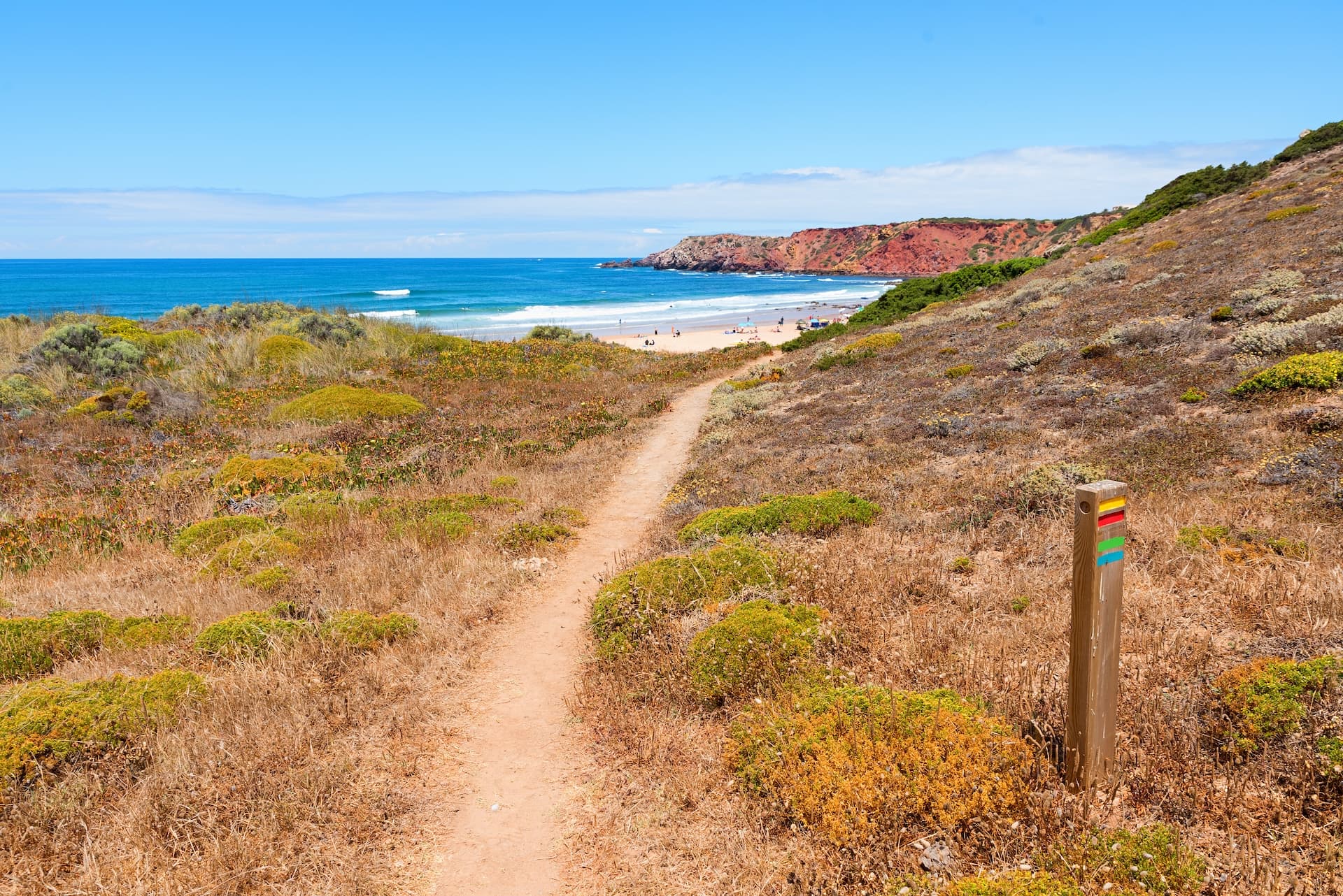 Hiking path along dry coastal scrub toward a beach with red cliffs in Algarve, Portugal.