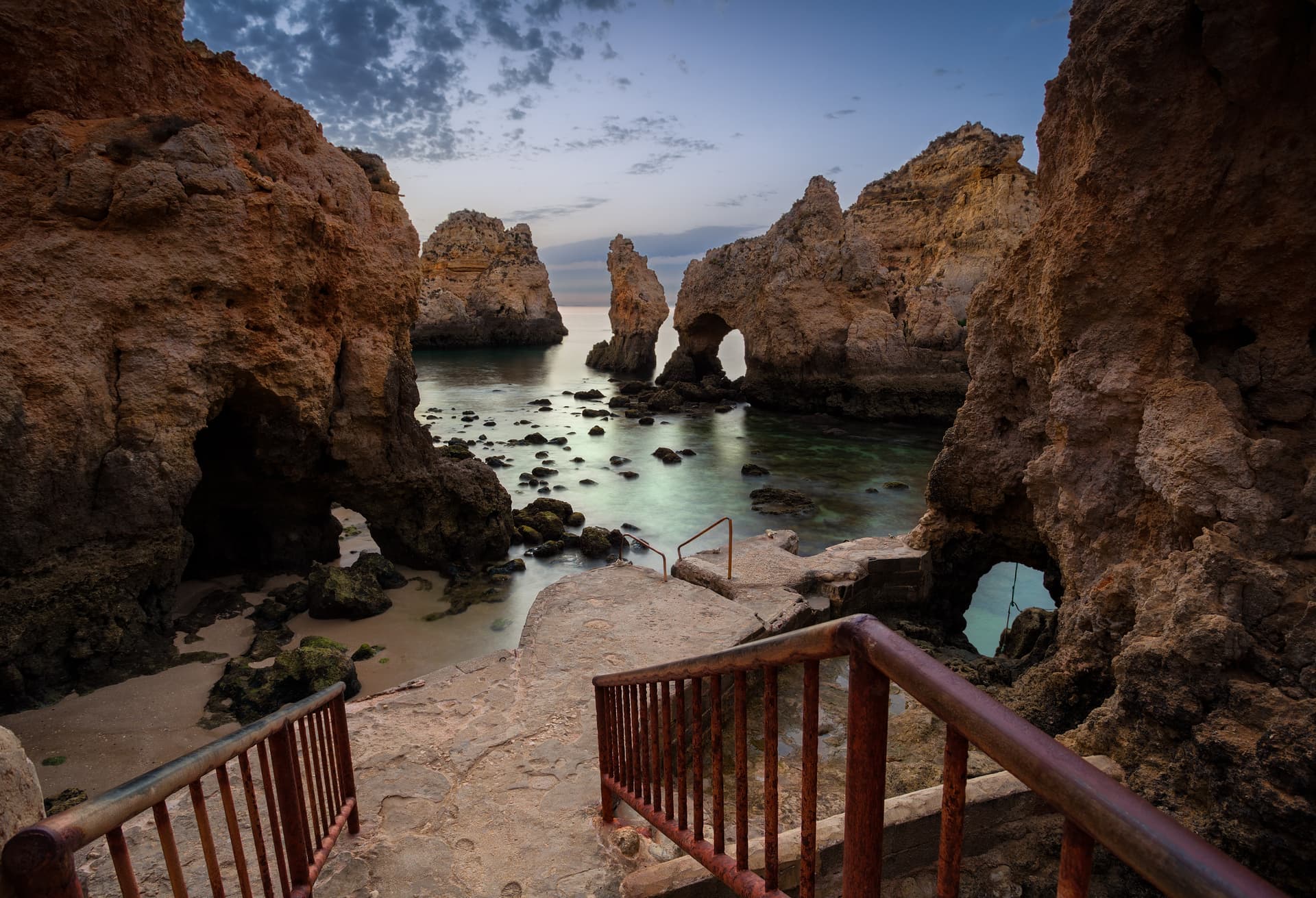 Stone steps with rusty railing leading to Ponta da Piedade sea caves at sunrise, Algarve, Portugal.