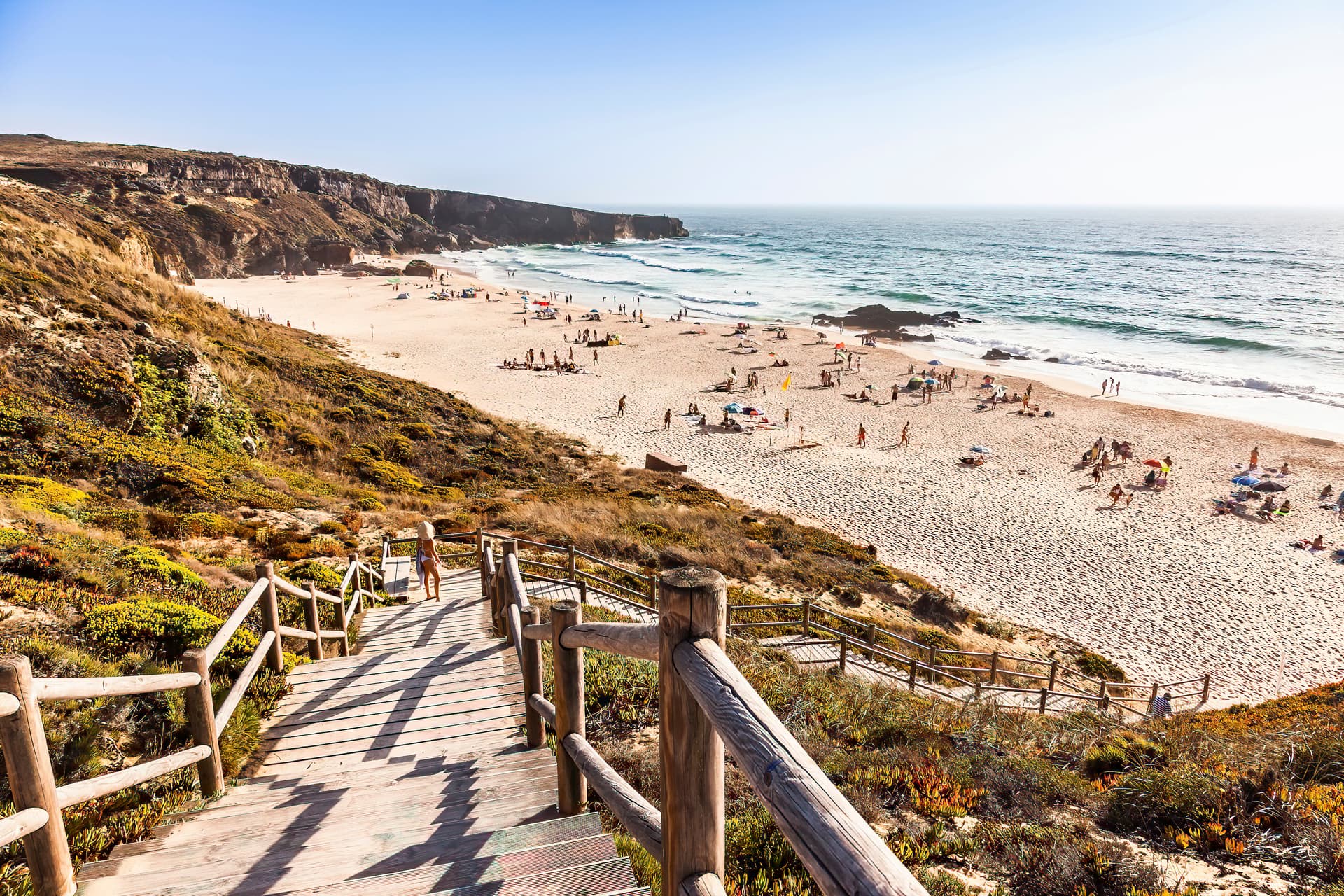 Wooden stairs lead down to a crowded beach with cliffs near Vila Nova de Milfontes, Alentejo, Portugal.