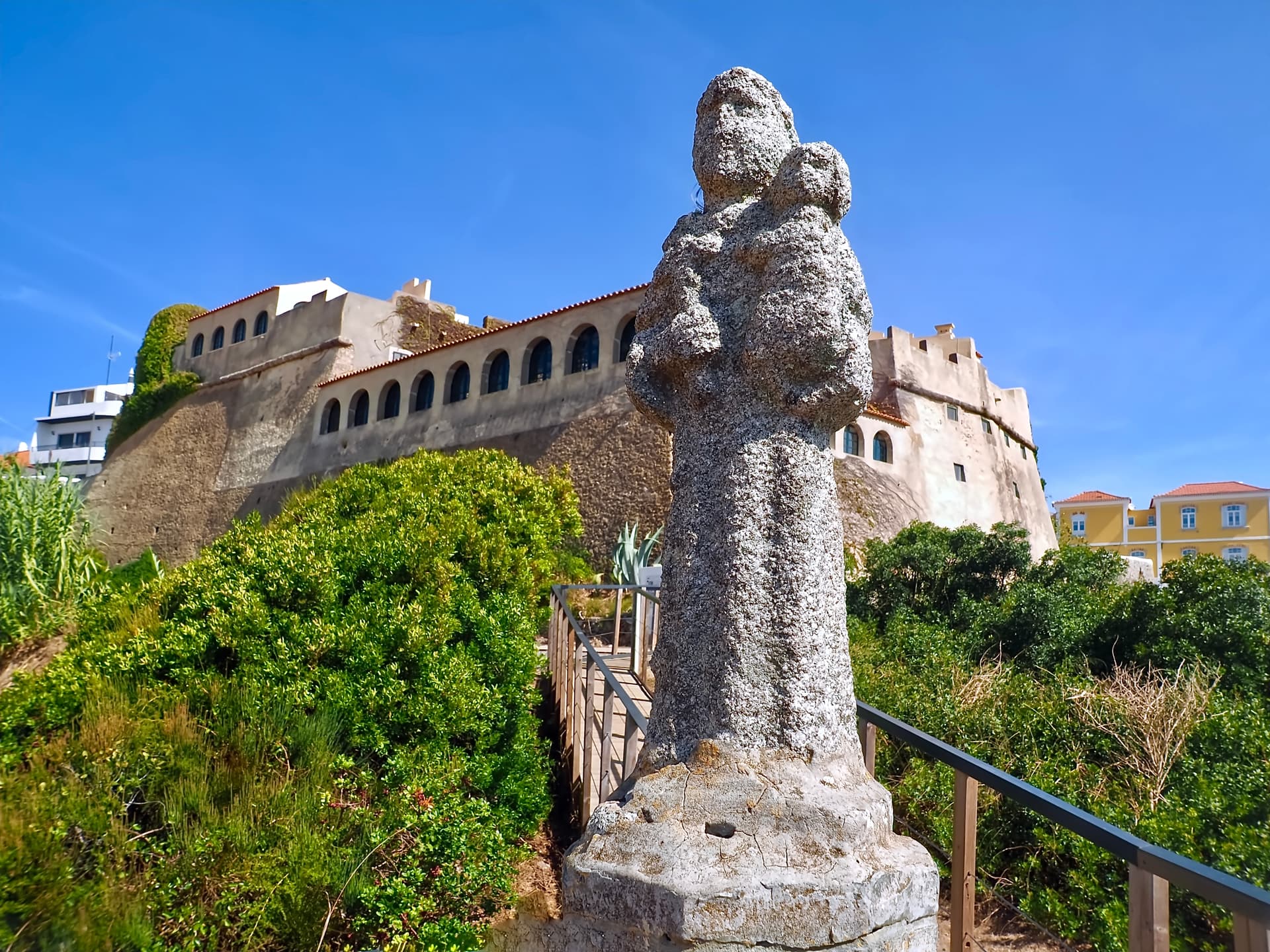 Stone sculpture with fort and green foliage under a clear blue sky in Vila Nova de Milfontes, Portugal.
