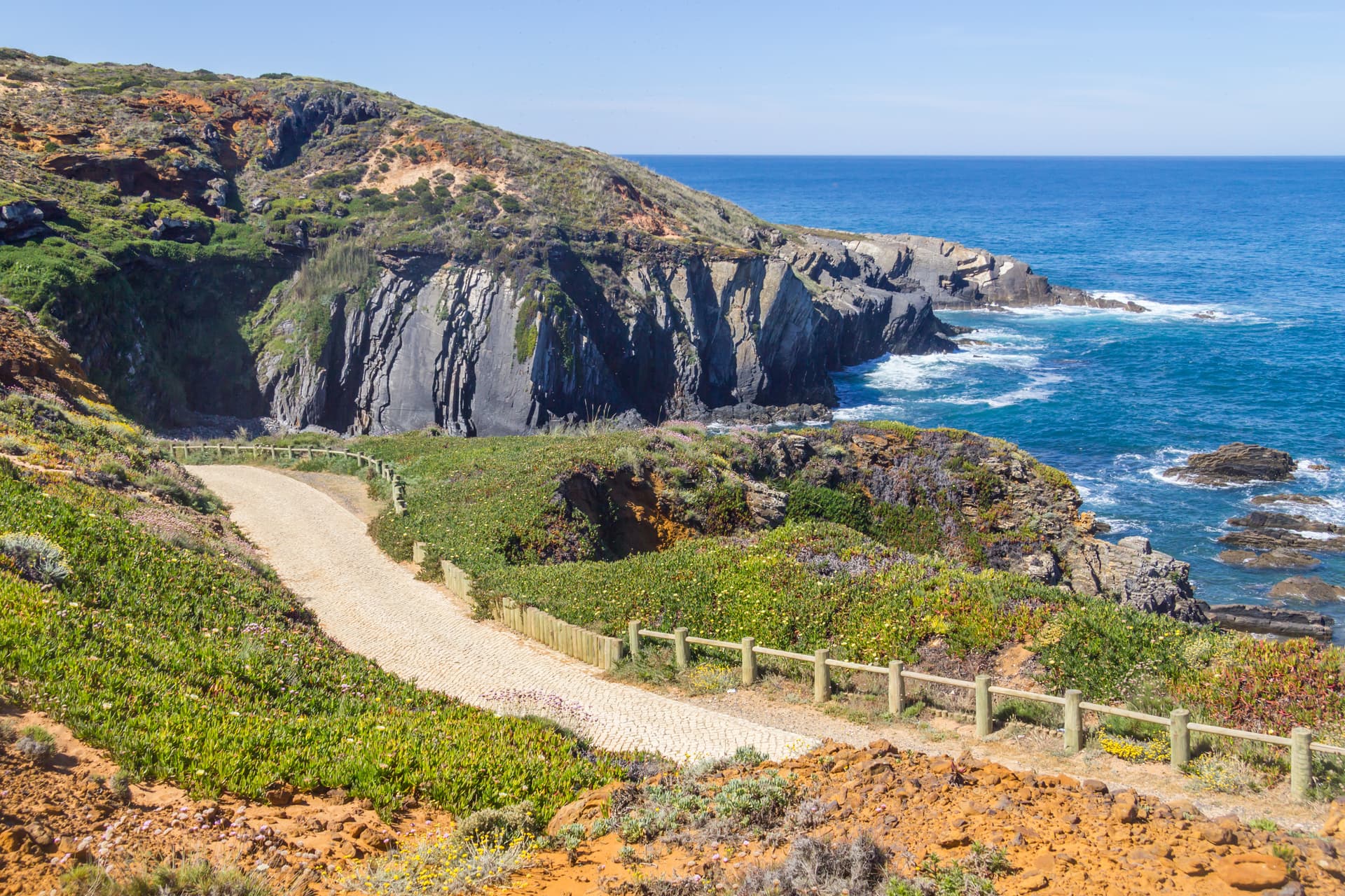 Trekking path along cliffs overlooking blue sea in Almograve with coastal vegetation.