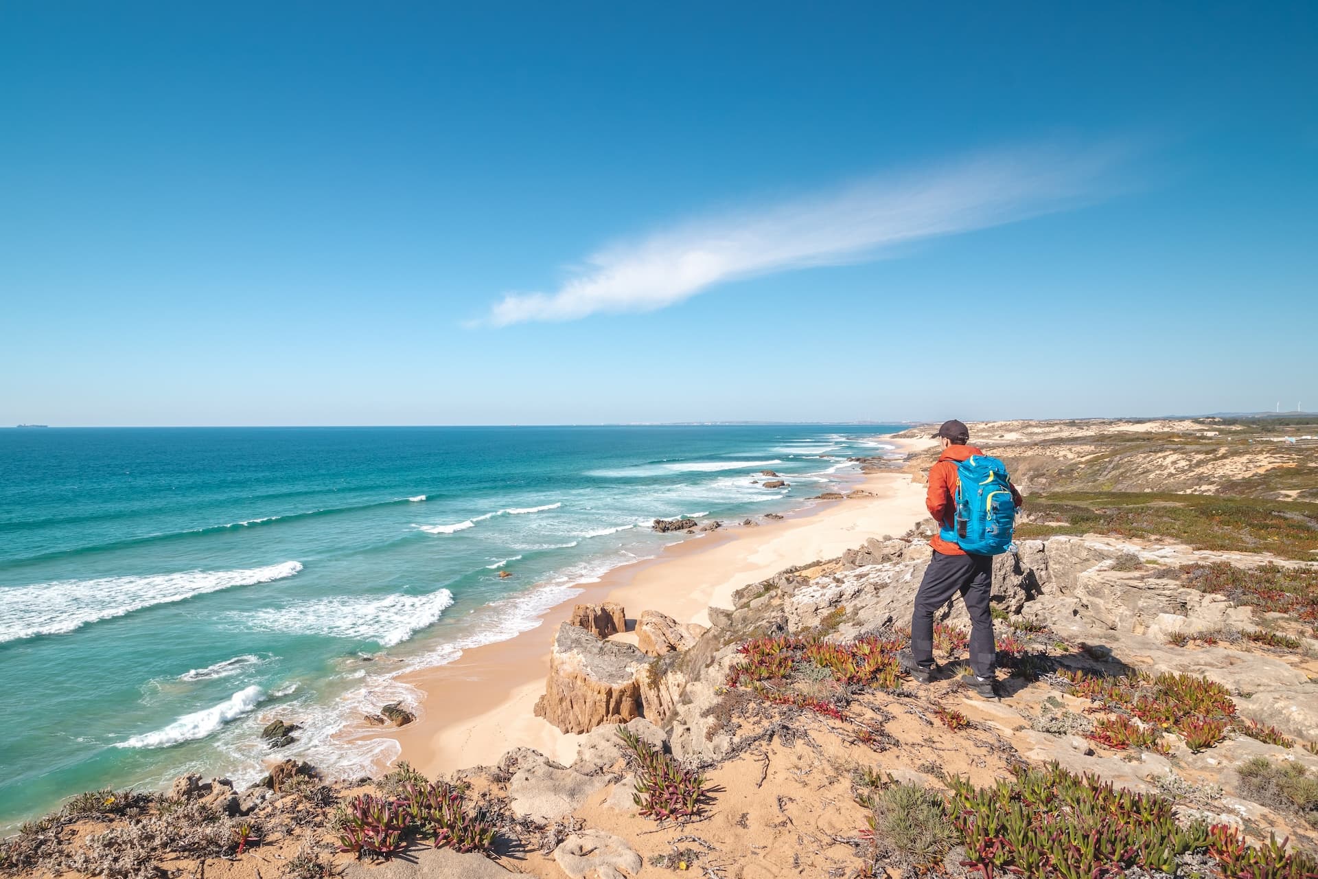 Hiker with blue backpack overlooking sandy beach and turquoise ocean near Vila Nova de Milfontes.