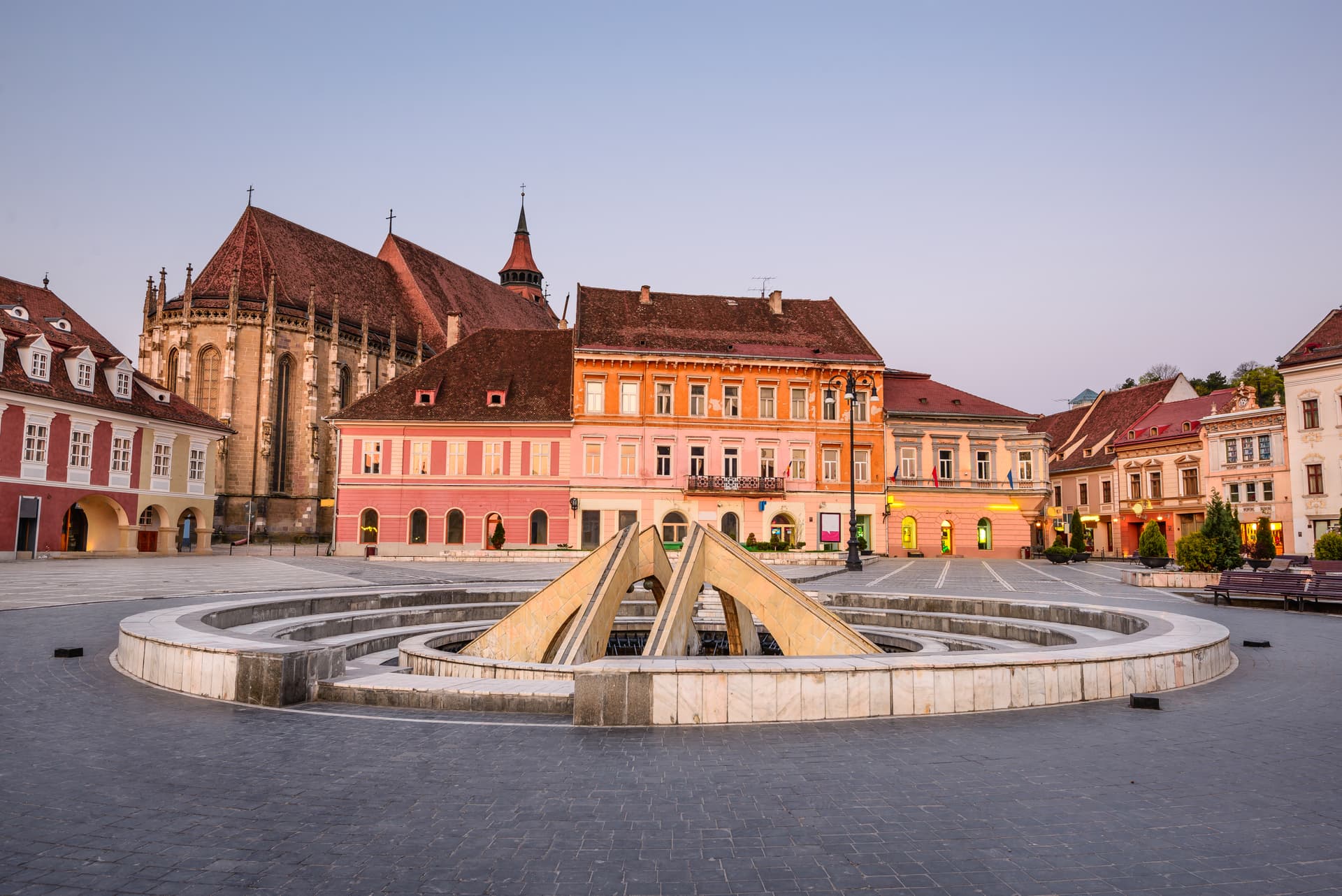 Historic buildings surround a circular fountain in Brasov Main Square at twilight.