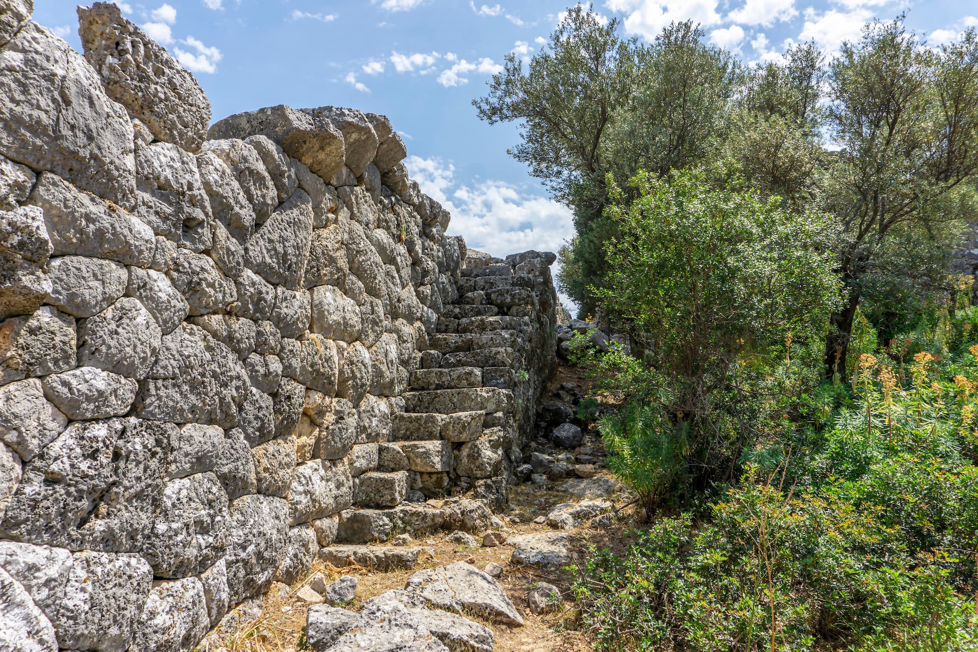Stone ruins wall and steps at Karadere under a partly cloudy blue sky with surrounding green foliage.