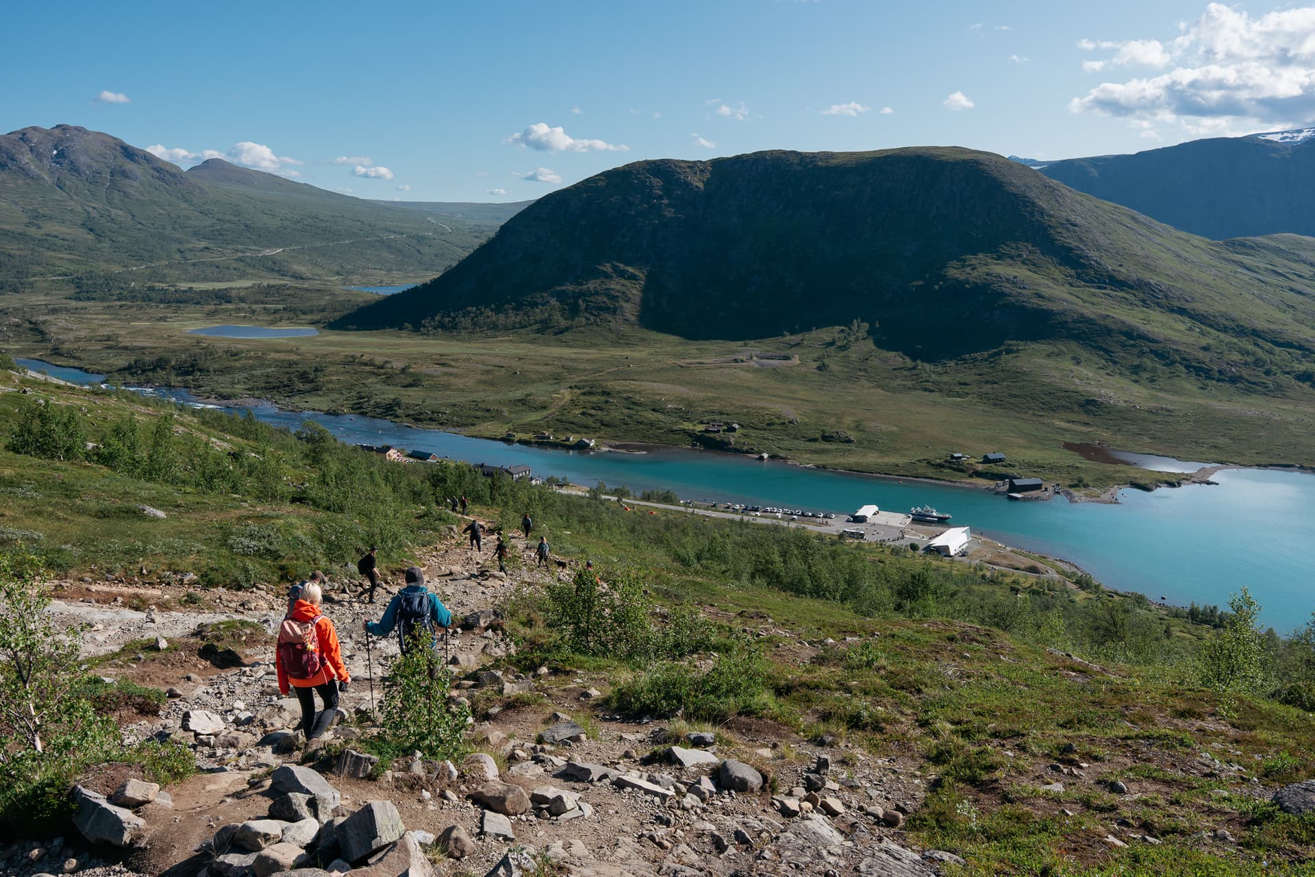 Hikers descending rocky trail toward Gjendesheim boat quay and turquoise lake in mountain valley