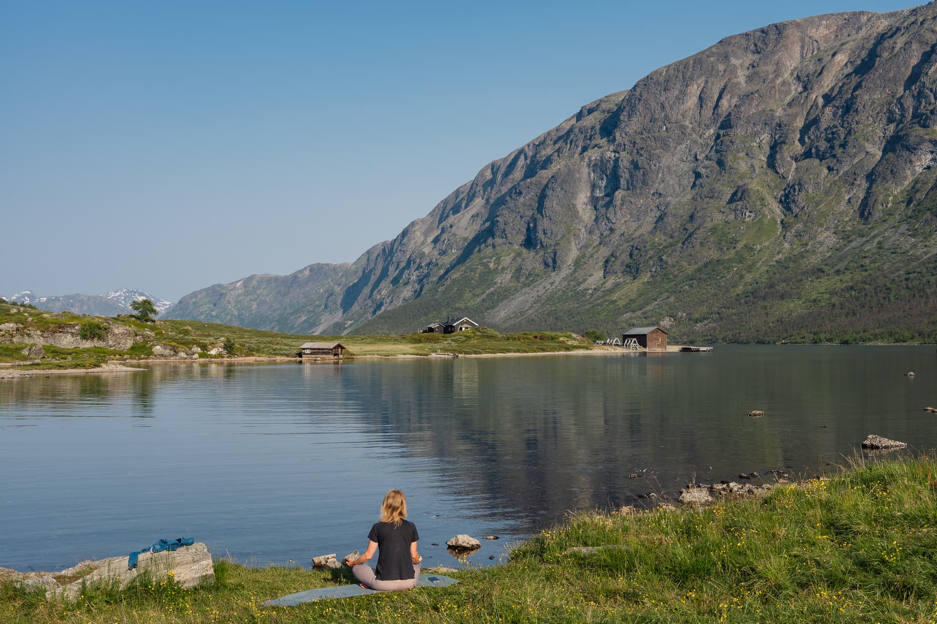 Person meditating on the shore of Lake Gjende with mountain backdrop and boathouses