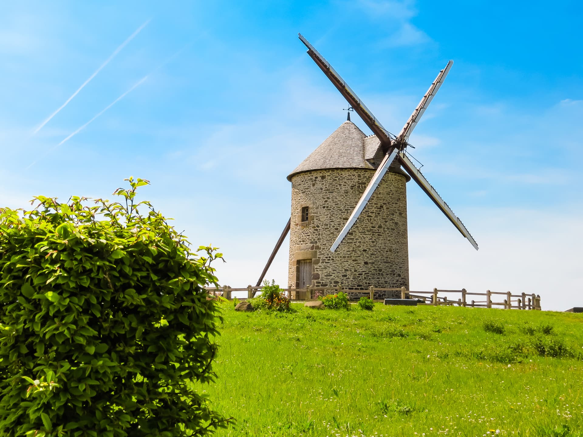 Stone windmill with wooden blades on a grassy hill under a bright blue sky in Normandy, France.