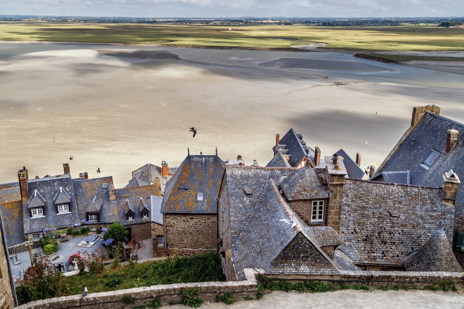 Mont Saint-Michel village rooftops overlooking low tide ocean flats and distant green fields.