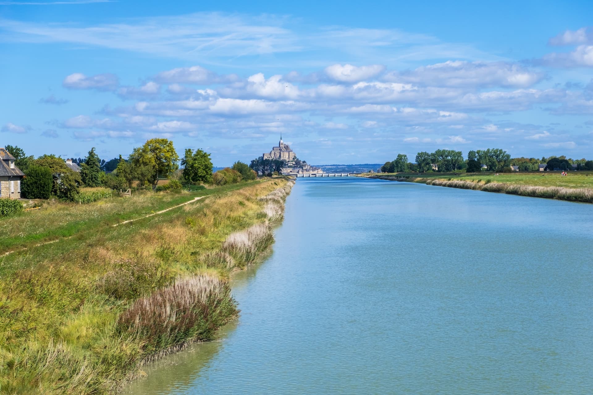 Couesnon River leads to Mont Saint-Michel abbey under a blue, cloudy sky in Normandy, France.