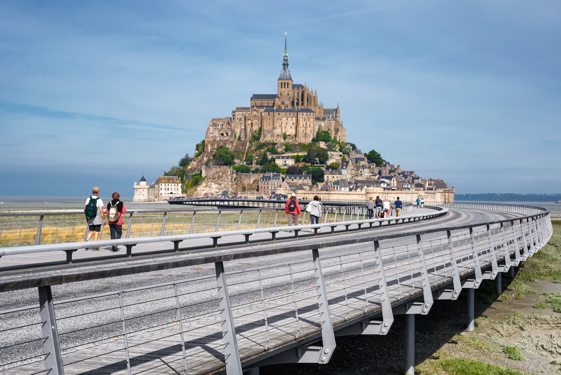 Tourists walking on bridge toward Mont Saint-Michel tidal island under blue sky.