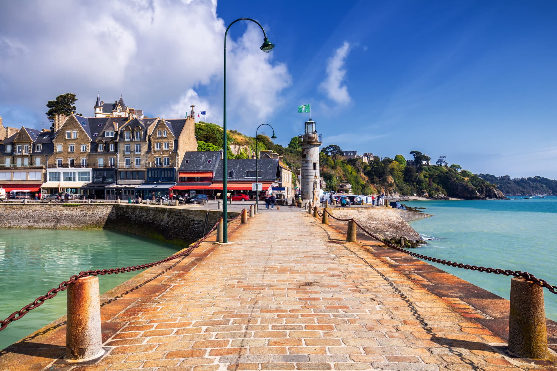 Brick pier leading to a stone lighthouse in Cancale, Brittany, with coastal buildings and green hills.