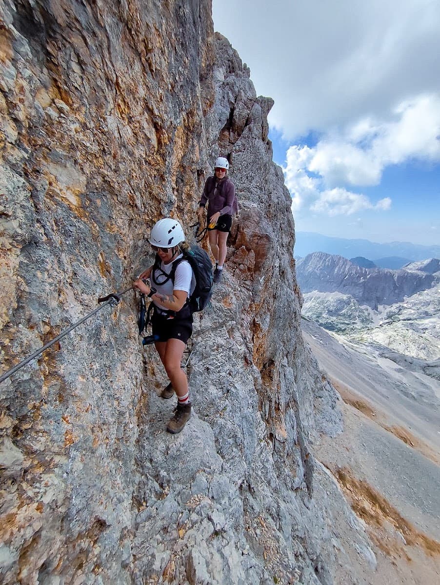 Two hikers with helmets and backpacks traversing a steep mountain ridge via a cable.
