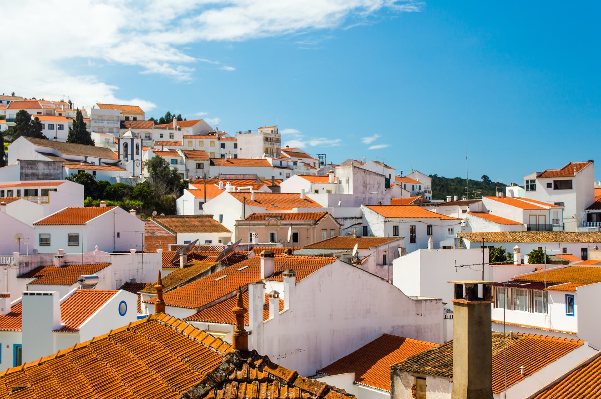 White houses with orange tile roofs clustered on a hillside under a bright blue sky in Odeceixe, Portugal.