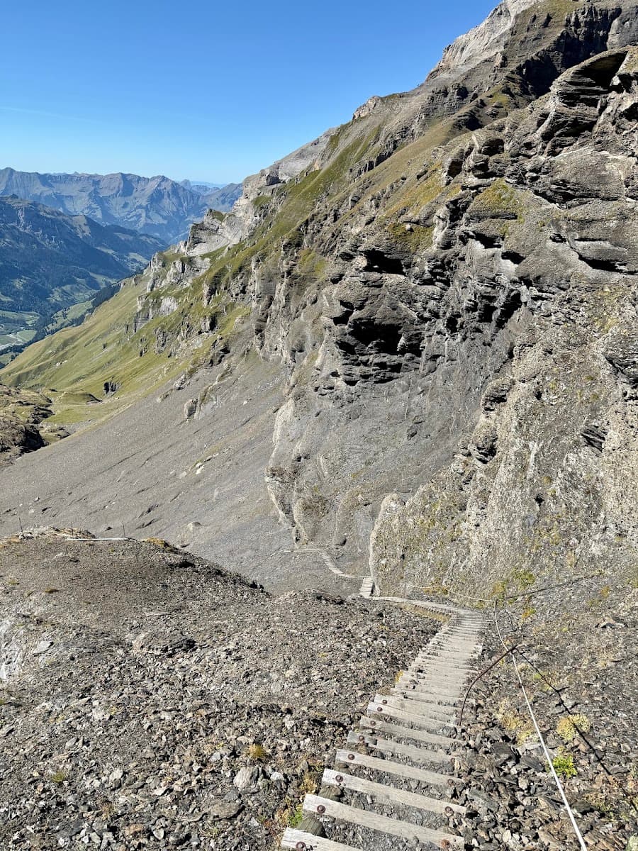 Steep wooden staircase descending along a rugged mountain cliffside with distant alpine peaks