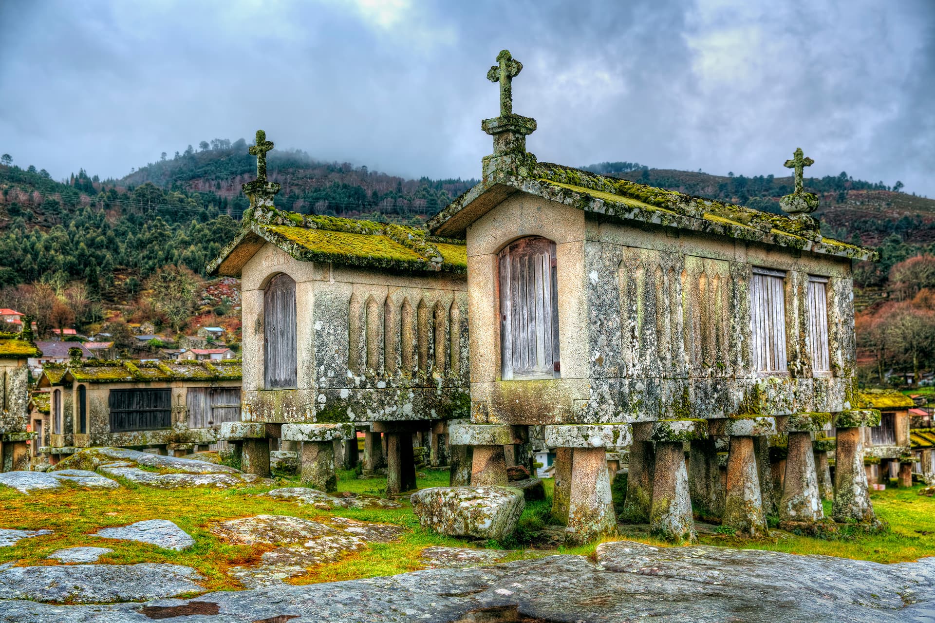 Moss-covered stone granaries with crosses on roofs in Lindoso, Portugal, against a forested hillside.