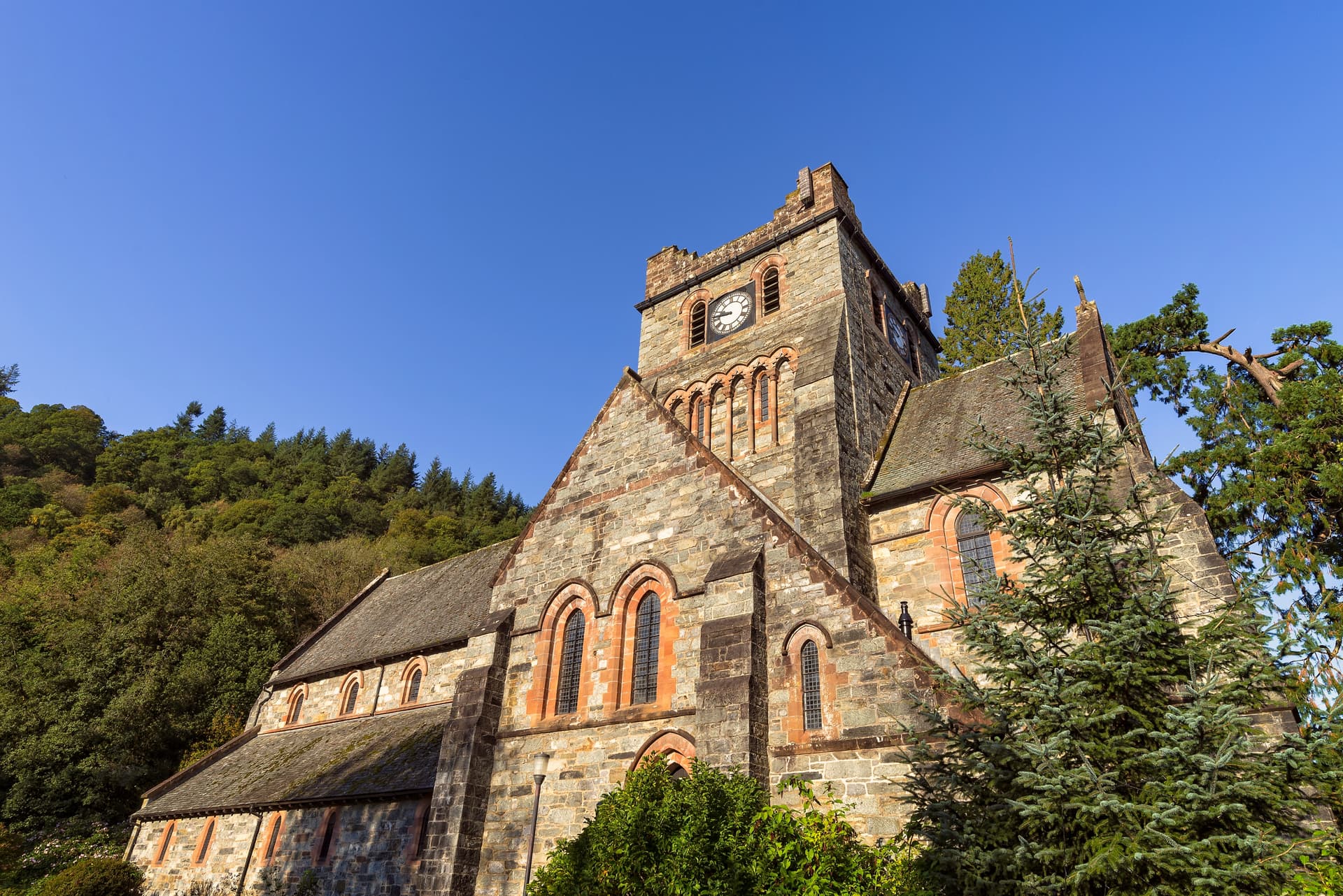 Stone church with clock tower against dense green hillside under clear blue sky in Betws-y-Coed.