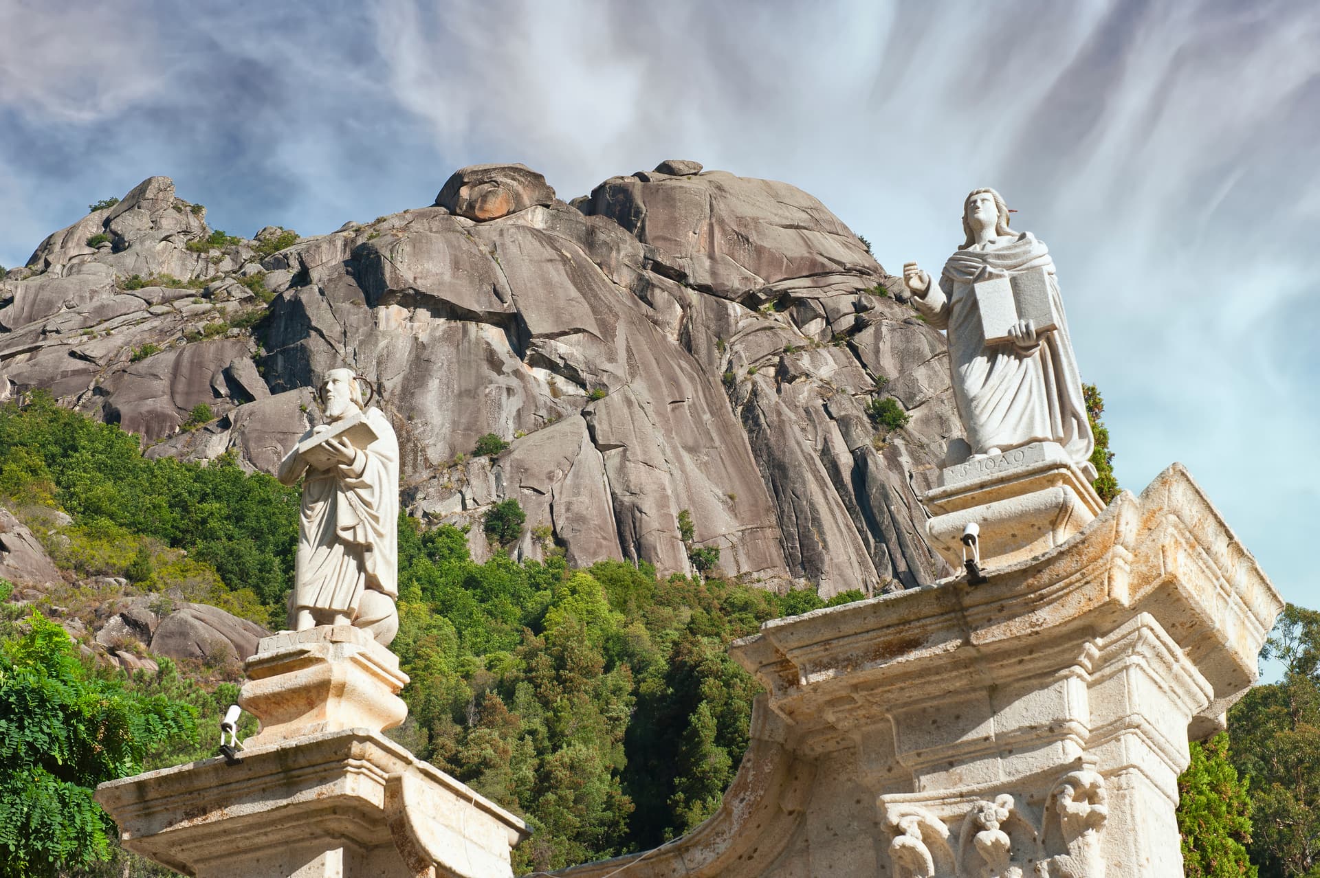 Stone statues flanking a massive granite mountain face with green forest below, Nossa Senhora da Peneda Sanctuary.