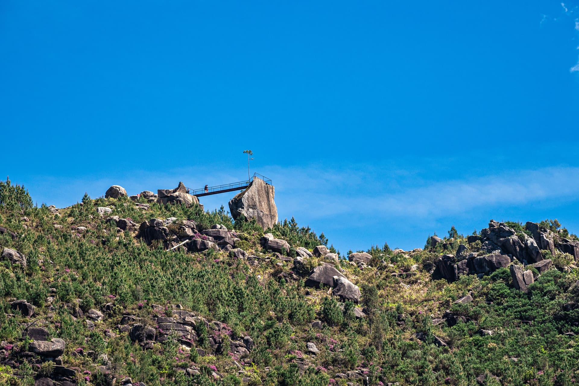 Viewpoint bridge connecting large rocks on a green, rocky hillside under a clear blue sky in Montalegre.