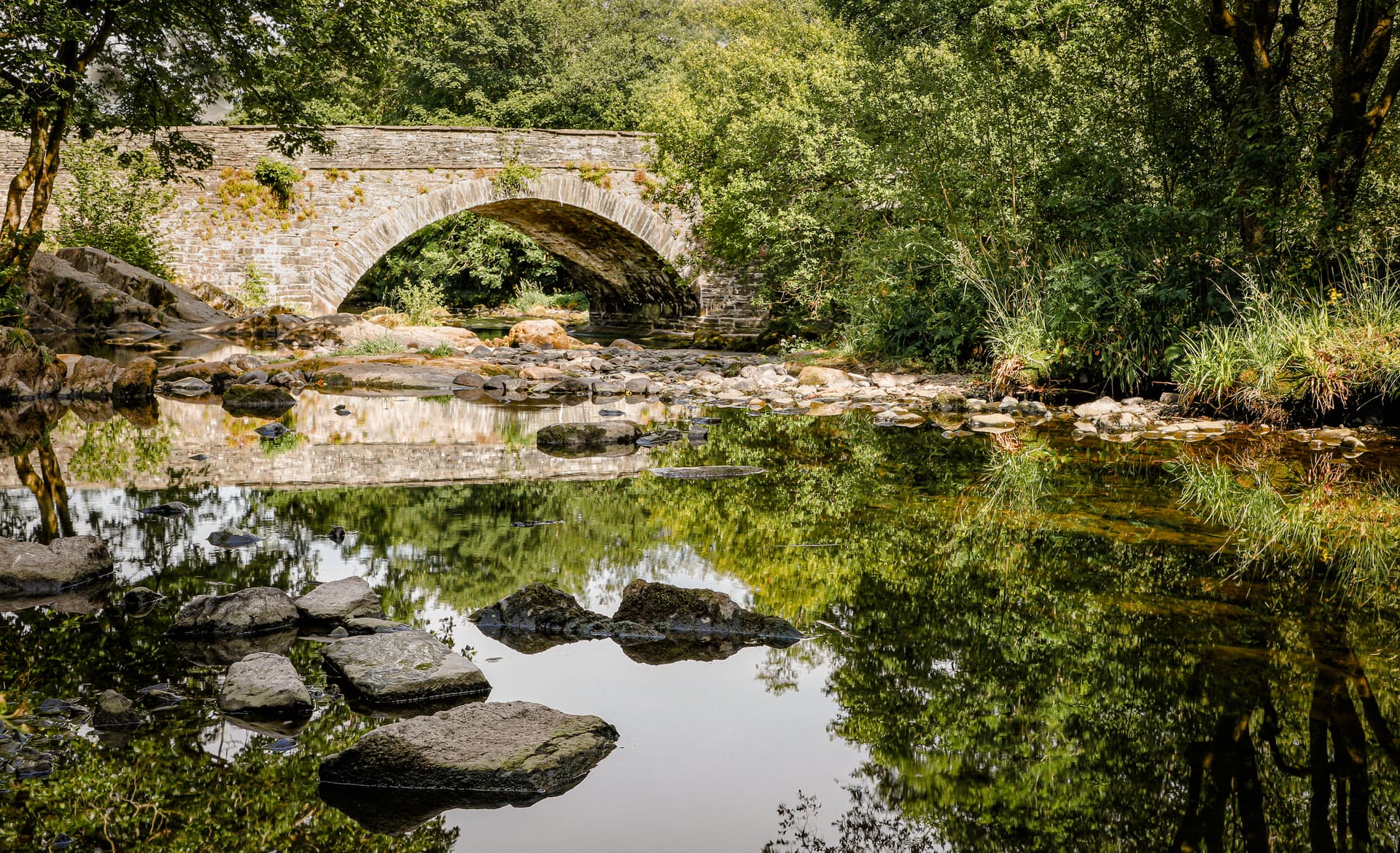Stone arch bridge over a rocky river with lush green trees and reflections in Ambleside.