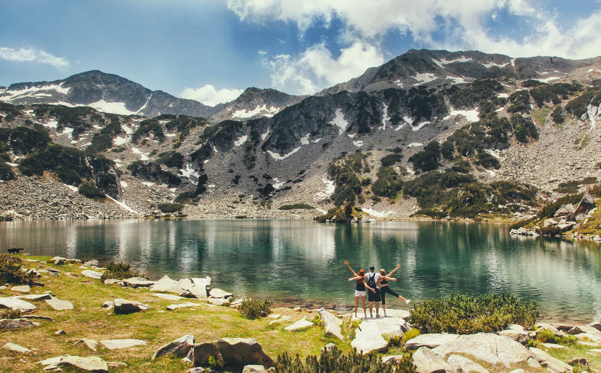 Hikers celebrating by clear alpine lake with rocky mountains and snow patches, Pirin Lakes