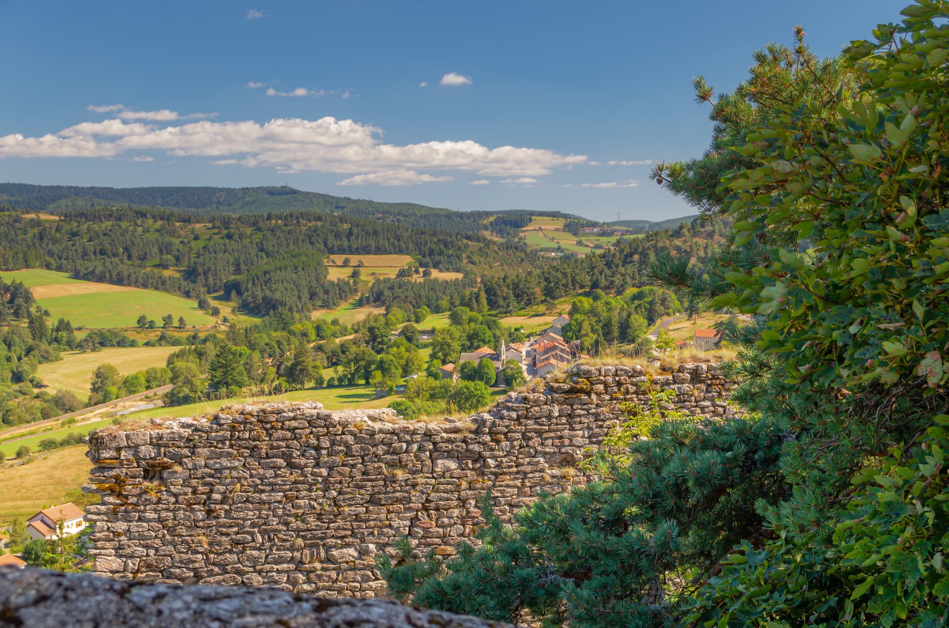 View from old stone wall over Lozerien landscape, rolling hills, forests, and village rooftops under blue sky.