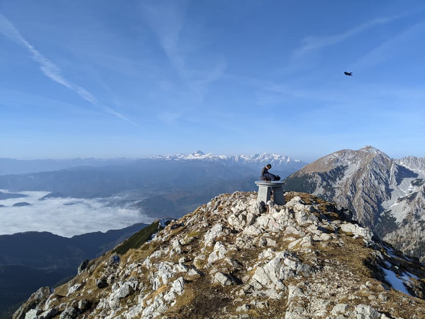 Hiker at stone summit marker on Begunjščica with view toward Stol and Julian Alps