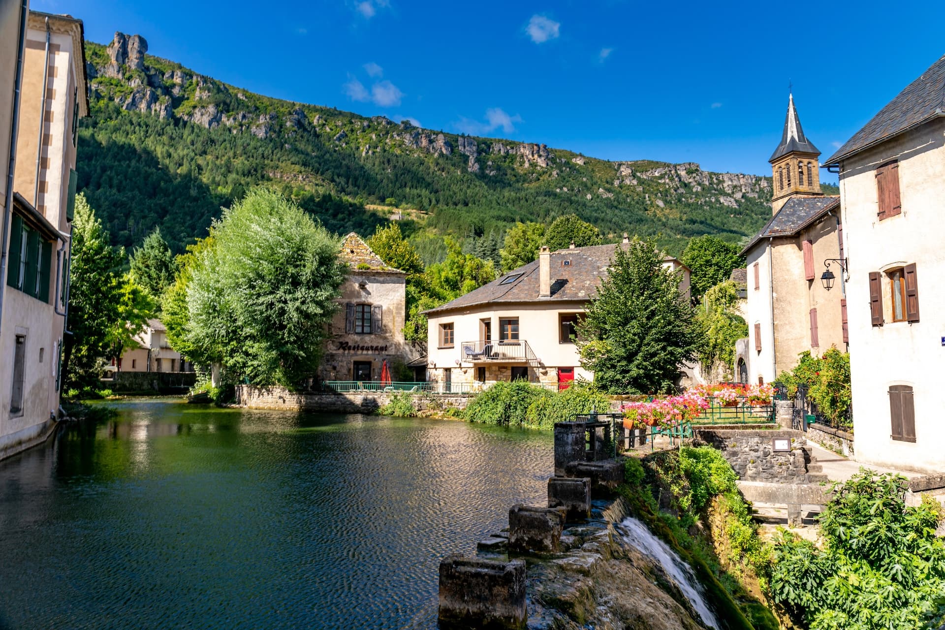Village of Florac Trois Rivières with river, stone buildings, and forested mountains under blue sky.