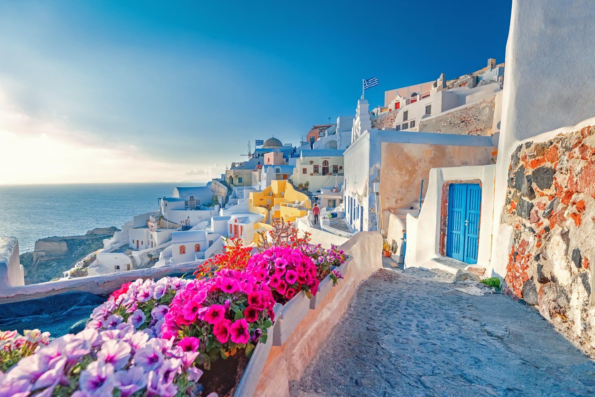 Santorini island street with white buildings, blue doors, and pink flowers overlooking the sea