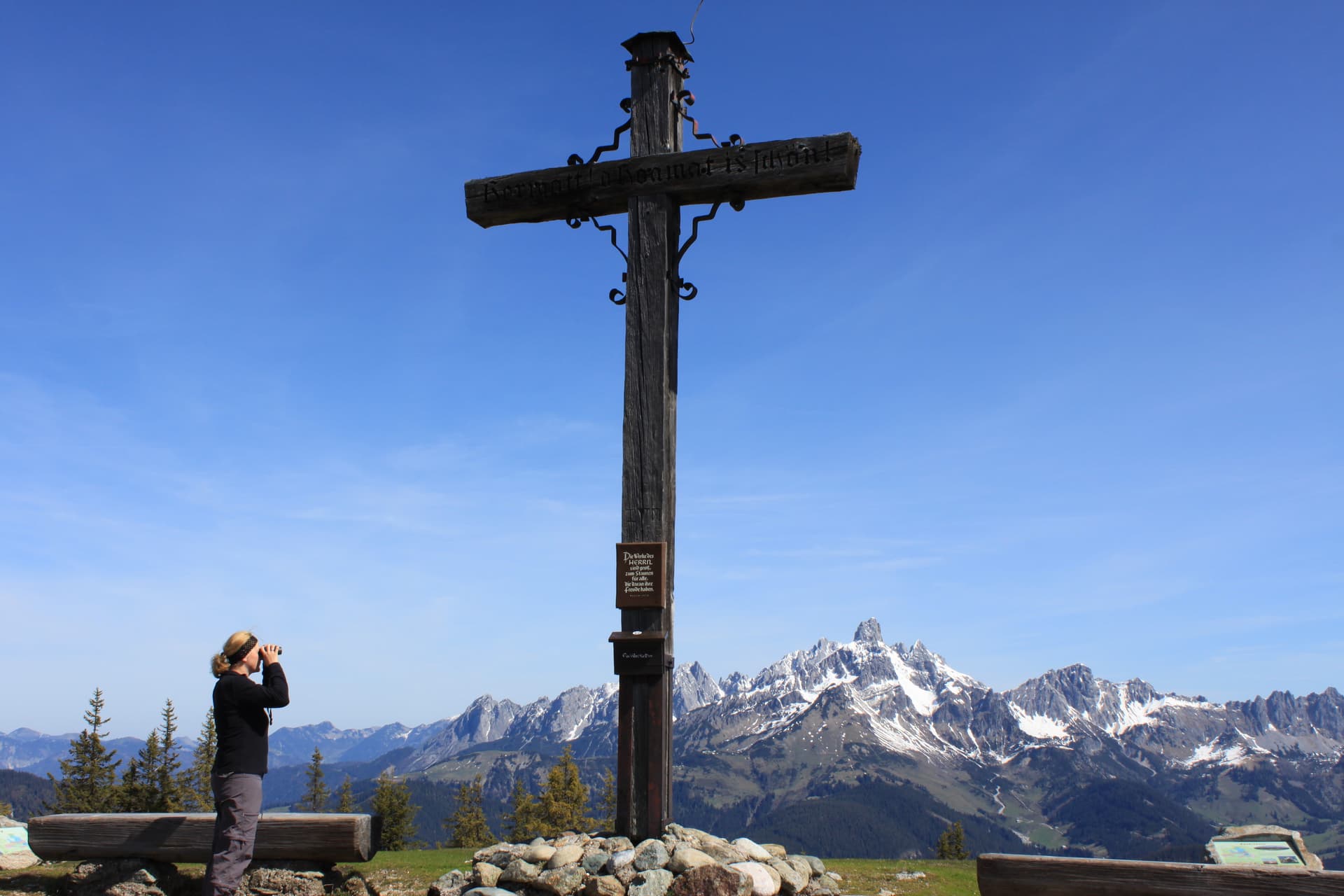 Hiker with binoculars by wooden summit cross overlooking snow-capped Rossbrand mountains.