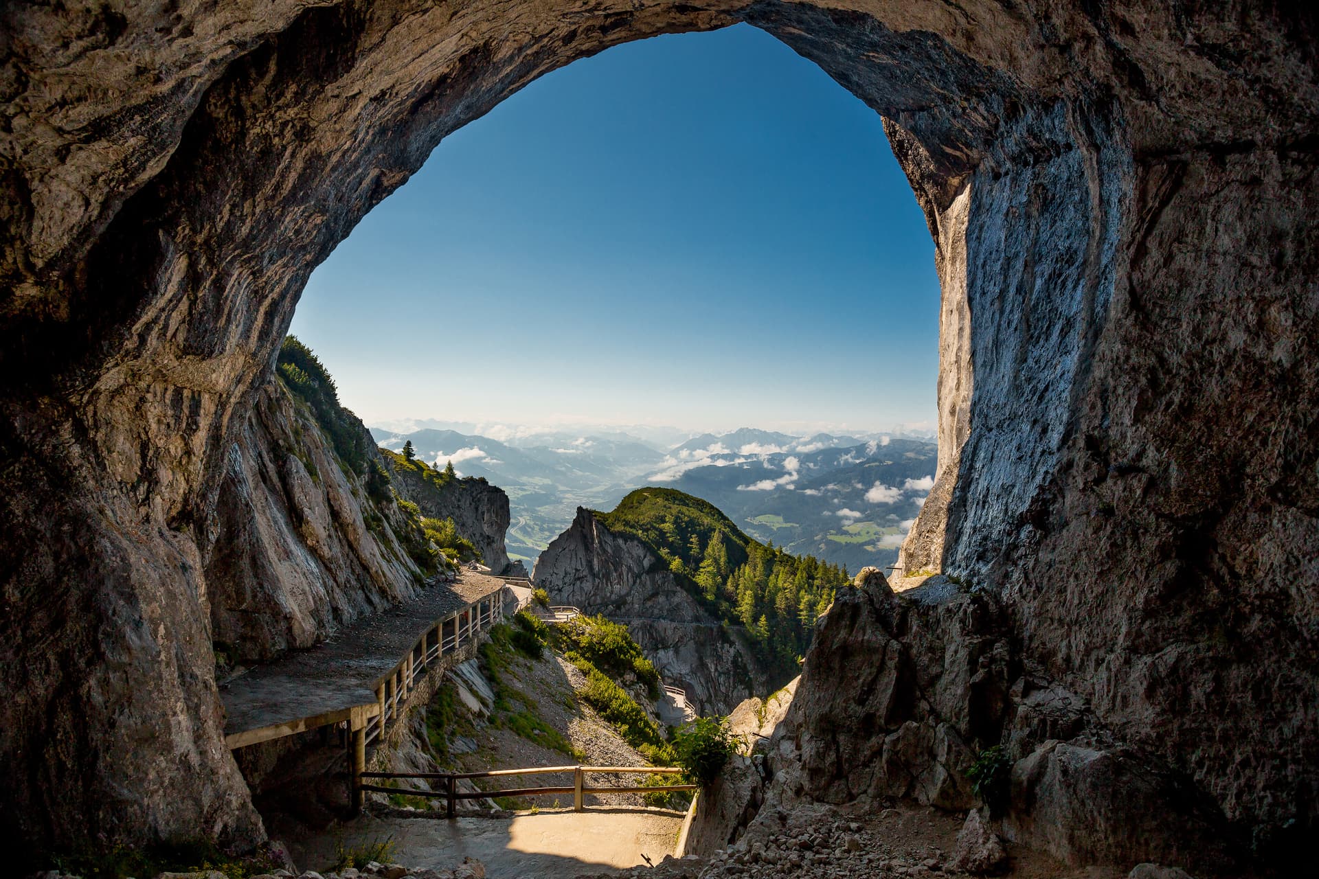 Cave opening framing a hiking path and view of green mountains under a clear blue sky.